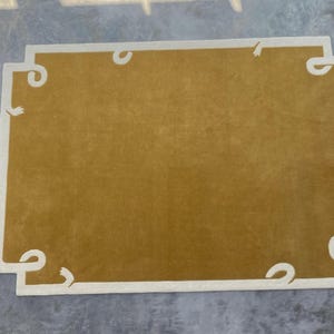 May include: Mustard-coloured rectangular rug with a white border and decorative scroll-like accents at each corner. The rug has a soft, plush texture. The rug is placed on a grey surface. The rug's dimensions are approximately 150cm x 200cm.