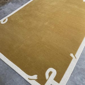 May include: Mustard yellow rectangular rug with a white border and decorative corner accents. The rug has a soft, textured appearance, suitable for a living room or bedroom. The design is modern and minimalist, adding a touch of elegance to any space.