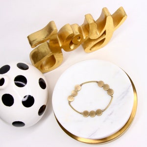 May include: A white ceramic vase with black polka dots, a gold-painted wooden sign that says "Joy", and a gold bracelet with round links on a white marble tray with a gold rim.