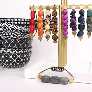 May include: A gold metal stand with eight bracelets made of various colorful stones and beads. The stand is on a white surface with a stack of black and white patterned bowls.