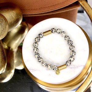 May include: A white and black speckled stone bracelet with gold accents. The bracelet is on a white marble surface.