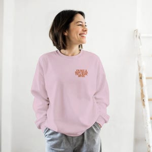 Könnte beinhalten: Hellrosa Sweatshirt mit dem orangefarbenen Text "I'M NOT A BOSS BITCH.". Das Sweatshirt mit Rundhalsausschnitt hat lange Ärmel. Die Person trägt eine graue Jogginghose.