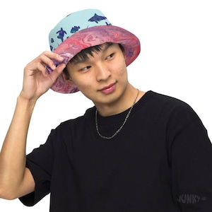 May include: A reversible bucket hat with a marine life design. The top of the hat is light blue with illustrations of sharks and turtles. The bottom half features a pink and purple marbled pattern. The hat is being held up by a person.