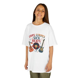 May include: White t-shirt featuring the text "Stars, Stripes & Sick Beats" and illustrations of a guitar, a record, and a guitar. The guitar and record have red, white, and blue accents. The shirt is worn by a person.