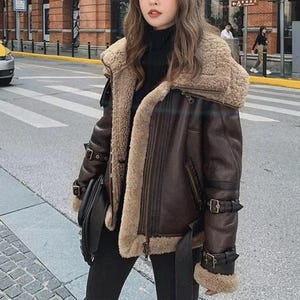 May include: A brown leather jacket with a shearling collar and lining. The jacket features multiple buckles, a belt, and a zip closure. The jacket is worn over a black turtleneck.