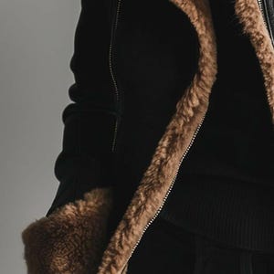 May include: Black jacket with brown faux fur trim and lining. The jacket has a zip closure and a ribbed knit cuff. The fur trim is visible along the collar, front, and cuffs, providing a contrasting texture and colour.