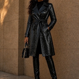 Könnte beinhalten: Ein schwarzer Trenchcoat aus Kunstleder mit Krokodilhautstruktur. Der Mantel hat ein doppelreihiges Design, einen Gürtel und Taschen. Das Model trägt schwarze Overknee-Stiefel und eine schwarze Handtasche.