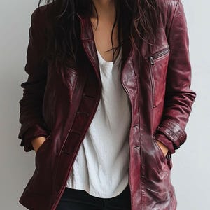 May include: A burgundy leather jacket with a button-up front and a zipped chest pocket. The jacket has a classic design with a notched collar and long sleeves. The model is wearing a white shirt underneath.
