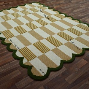 Puede incluir: Una alfombra rectangular con un dise&ntilde;o a cuadros en beige y blanco, bordeada por un borde festoneado en verde oscuro. La alfombra est&aacute; colocada sobre un suelo de madera. El texto "Art of rug india" es visible.