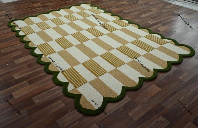 Puede incluir: Una alfombra rectangular con un dise&ntilde;o a cuadros en beige y blanco, bordeada por un borde festoneado en verde oscuro. La alfombra est&aacute; colocada sobre un suelo de madera. El texto "Art of rug india" es visible.