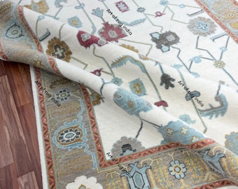 May include: Ivory-colored rug with a detailed floral pattern in shades of blue, red, and brown. The border features a repeating geometric design with a touch of gold. The rug is partially rolled up, revealing its intricate weave. The text "Art of rug india" is visible.