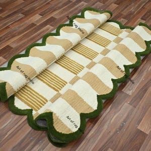 Puede incluir: Una alfombra rectangular con un borde verde festoneado. La alfombra presenta un patr&oacute;n de rayas y formas beige y blancas alternas. La alfombra est&aacute; parcialmente enrollada, revelando la parte inferior. La alfombra est&aacute; sobre un suelo de madera.