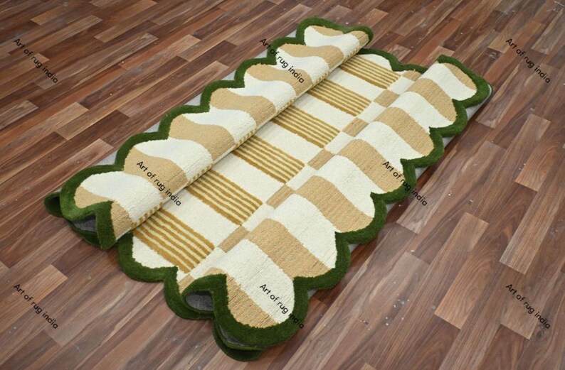 Puede incluir: Una alfombra rectangular con un borde verde festoneado. La alfombra presenta un patr&oacute;n de rayas y formas beige y blancas alternas. La alfombra est&aacute; parcialmente enrollada, revelando la parte inferior. La alfombra est&aacute; sobre un suelo de madera.