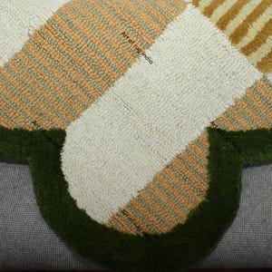 Puede incluir: Primer plano de una alfombra hecha a mano con forma de tr&eacute;bol. La alfombra presenta un patr&oacute;n de secciones crema, naranja y beige, bordeado por un borde verde oscuro. El texto "Art of rug india" es visible.