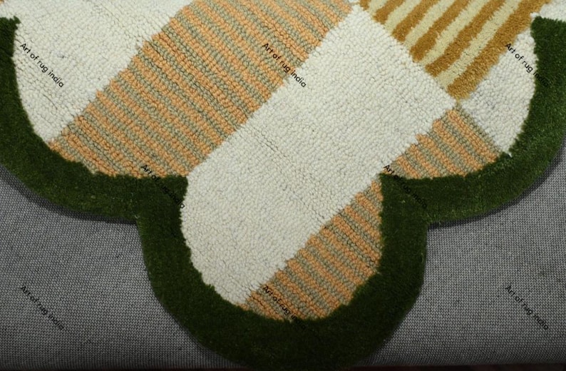 Puede incluir: Primer plano de una alfombra hecha a mano con forma de tr&eacute;bol. La alfombra presenta un patr&oacute;n de secciones crema, naranja y beige, bordeado por un borde verde oscuro. El texto "Art of rug india" es visible.
