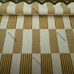 Puede incluir: Una alfombra a rayas enrollada con rayas verticales beige y blancas alternas. La alfombra tiene un borde verde festoneado. El texto "Art of rug india" es visible en la alfombra.