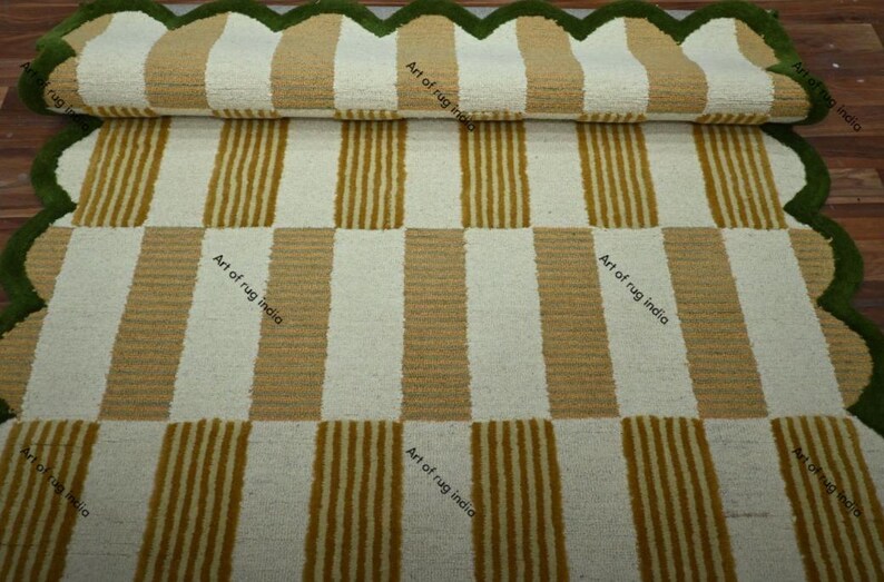 Puede incluir: Una alfombra a rayas enrollada con rayas verticales beige y blancas alternas. La alfombra tiene un borde verde festoneado. El texto "Art of rug india" es visible en la alfombra.