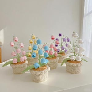 Op de afbeelding: Een verzameling handgemaakte gehaakte bloemstukken in kleine potten. Elke pot heeft bloemen in verschillende kleuren, waaronder roze, geel, blauw, paars en wit. De potten zijn beige met een geschulpte rand.