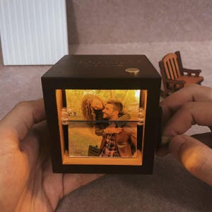 Puede incluir: Un cubo de fotos de madera oscura con una foto de una pareja abrazándose, iluminada desde dentro. El cubo tiene un pequeño botón dorado en la parte superior y un mecanismo giratorio en el lateral. La imagen se muestra con una luz cálida.
