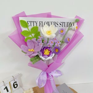 Op de afbeelding: Een boeket gehaakte bloemen in paars, wit en geel, verpakt in lavendelkleurig papier en vastgebonden met een bijpassend lint. Het boeket bevat verschillende bloemsoorten en groene bladeren. De tekst "ETTY FLOWERS STUDIO" is zichtbaar.