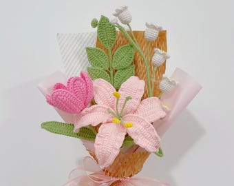 May include: A handmade crochet bouquet featuring a pink lily, a pink tulip, and white bell-shaped flowers. The bouquet is wrapped in pink paper and tied with a pink ribbon. Green leaves and stems add to the floral arrangement.