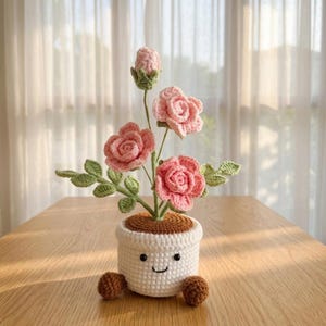 Puede incluir: Una planta de ganchillo en maceta con tres rosas rosadas y un capullo cerrado sobre tallos verdes con hojas. La maceta blanca tiene una parte superior marrón, dos pies pequeños marrones y una cara bordada con dos ojos negros y una boca.
