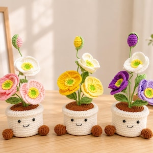 Op de afbeelding: Drie gehaakte bloemen in lachende witte potten. De linkerpot heeft roze en witte bloemen, de middelste gele en witte bloemen en de rechter paarse en witte bloemen. Elke plant is voorzien van groene bladeren en kleine bloemknoppen.