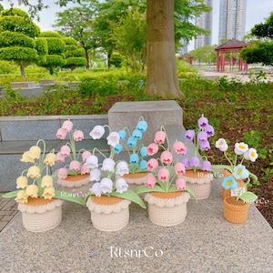 May include: A collection of handmade crochet flower arrangements in various colors, including yellow, pink, white, blue, and purple. Each arrangement is in a beige pot with a scalloped edge, creating a charming, decorative display.