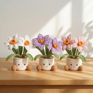 May include: Three crocheted daffodil plants in white, purple, and pink, each with an orange centre and green leaves. The flowers are in small white pots with smiling faces and brown feet, displayed on a wooden surface.