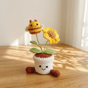Può includere: Una pianta in vaso all'uncinetto con un'ape a strisce gialle e marroni e un girasole giallo con il centro marrone. Il vaso bianco all'uncinetto ha un volto sorridente con occhi a perla neri e due piccoli piedi marroni alla base del vaso.