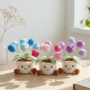 May include: Three crocheted potted plants featuring colorful tulip-shaped flowers in shades of blue, pink, and purple. Each white pot has a stitched smiling face, two small brown feet, and green crocheted leaves supporting the yarn flowers.
