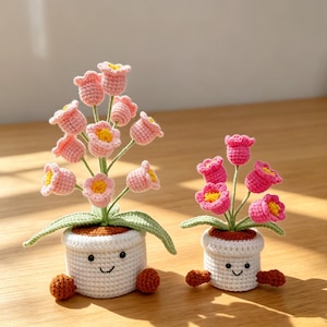 May include: Two crochet flower arrangements in white pots with smiling faces. The flowers are pink with yellow centres and green leaves. The pots have brown feet and are on a wooden surface.