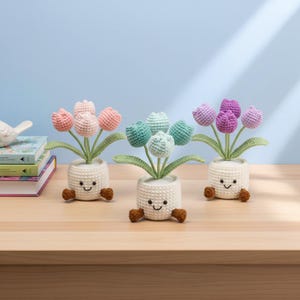 Op de afbeelding: Drie gehaakte tulpenplanten in witte potten met lachende gezichten en bruine voetjes. De tulpen zijn in tinten roze, blauwgroen en paars. De planten staan op een houten oppervlak met een blauwe achtergrond.