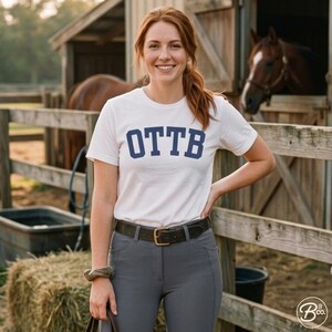 May include: White t-shirt with the navy blue letters OTTB. The person is wearing gray pants and a brown belt with a gold buckle. A horse is visible in the background, along with a wooden fence and a barn.