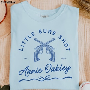 May include: Light blue t-shirt with a navy blue graphic featuring crossed revolvers and the text "LITTLE SURE SHOT" and "Annie Oakley". The shirt also includes the text "EST. 1860".