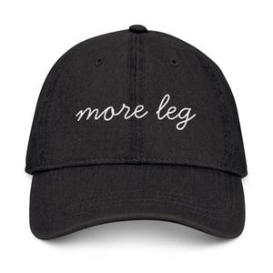 Könnte beinhalten: Schwarze Baseballkappe mit dem gestickten Text "more leg" in weißer Schrift. Die Kappe hat einen gebogenen Schirm und einen Knopf oben. Ein lässiges Accessoire.