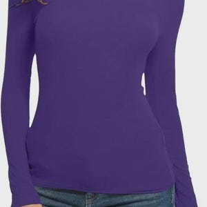 Pu&ograve; includere: Una maglia viola a maniche lunghe con collo alto. La maglia &egrave; di colore unito e sembra essere realizzata in materiale elasticizzato. La modella indossa anche jeans blu.