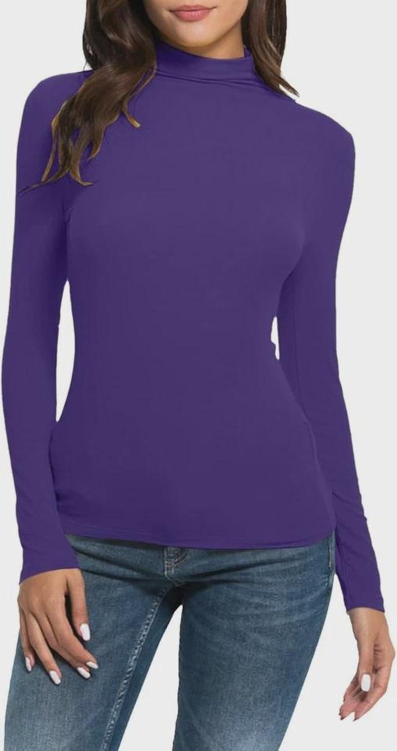 Pu&ograve; includere: Una maglia viola a maniche lunghe con collo alto. La maglia &egrave; di colore unito e sembra essere realizzata in materiale elasticizzato. La modella indossa anche jeans blu.