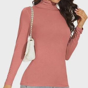 Pu&ograve; includere: Una maglia a maniche lunghe con collo alto rosa polveroso. La maglia &egrave; aderente ed &egrave; realizzata in un materiale morbido. Una borsa a tracolla bianca trapuntata con una tracolla a catena dorata &egrave; indossata sulla spalla. La persona indossa jeans blu.
