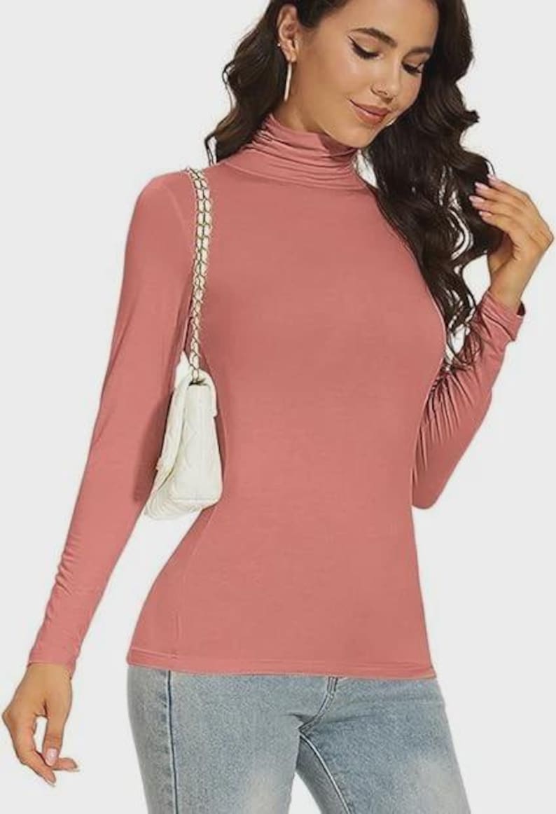 Pu&ograve; includere: Una maglia a maniche lunghe con collo alto rosa polveroso. La maglia &egrave; aderente ed &egrave; realizzata in un materiale morbido. Una borsa a tracolla bianca trapuntata con una tracolla a catena dorata &egrave; indossata sulla spalla. La persona indossa jeans blu.