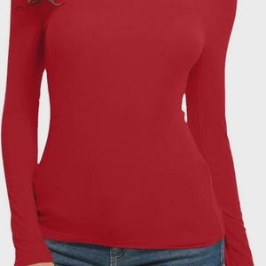 Pu&ograve; includere: Una maglietta rossa a maniche lunghe con collo alto. La maglietta &egrave; di colore unito e sembra essere fatta di un materiale morbido ed elastico. La modella indossa jeans blu.