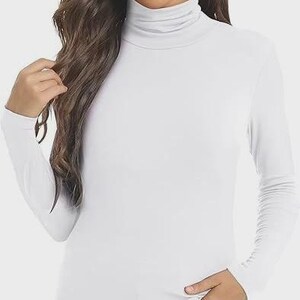 Pu&ograve; includere: Una maglietta bianca a maniche lunghe con collo alto. La maglietta &egrave; realizzata in un materiale morbido ed elastico ed &egrave; abbinata a jeans blu. La modella indossa la maglietta e i jeans.