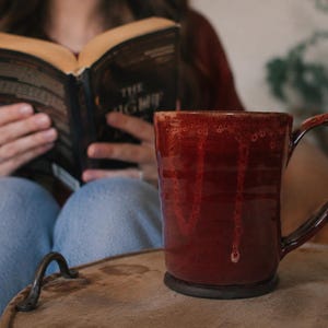 May include: A deep red ceramic mug with a glossy, dripped glaze finish and a curved handle. The mug features a dark, solid base and a textured, uneven rim. The rich, dark red color transitions into lighter, translucent drips down the sides.