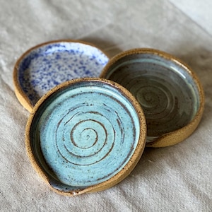 May include: Three handmade ceramic spoon rests. The spoon rests are round with a shallow dish and a small notch. One is light blue with a spiral design, one is blue with white speckles, and the third is a muted green with a spiral design.