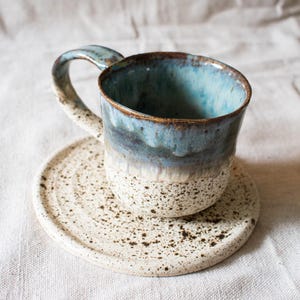 May include: A ceramic mug and matching saucer set featuring a speckled cream base with dark brown flecks. The upper portion of the mug and the handle are finished in a glossy blue glaze that transitions into the speckled cream texture below.