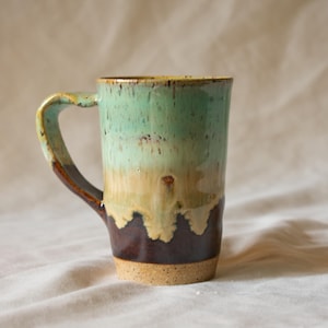 May include: A ceramic mug featuring a vertical gradient glaze that transitions from a speckled mint green at the top to a tan middle section and a dark brown base. The mug has a single curved handle and a textured, unglazed speckled tan bottom rim.