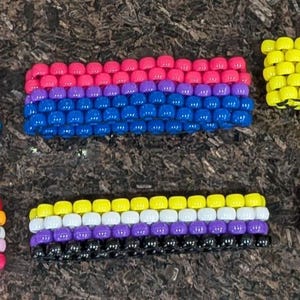 May include: Six rectangular bracelets crafted from small plastic pony beads arranged in distinct horizontal color patterns. The bracelets feature various color combinations including pink, white, blue, purple, yellow, orange, and black in striped designs.