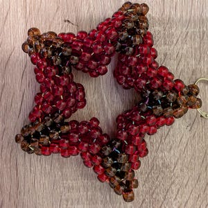 May include: A star-shaped keychain made of red and brown beads. The beaded star has a silver-colored keyring attached. The beads are a mix of red and brown, creating a textured appearance. The keychain is on a light brown wooden surface.