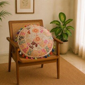 Könnte beinhalten: Ein runder Patchwork-Kissen mit verschiedenen bunten Mustern und Quasten liegt auf einem Holzstuhl. Das Kissen zeigt florale und geometrische Designs in Rosa, Orange und Blau. Der Stuhl hat eine gewebte Rückenlehne und Armlehnen.
