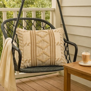 Puede incluir: Un cojín decorativo beige y marrón con un patrón geométrico descansa sobre una silla colgante gris oscuro. Una manta de color crema está sobre el brazo de la silla, y una pequeña mesa de madera contiene un vaso de café.